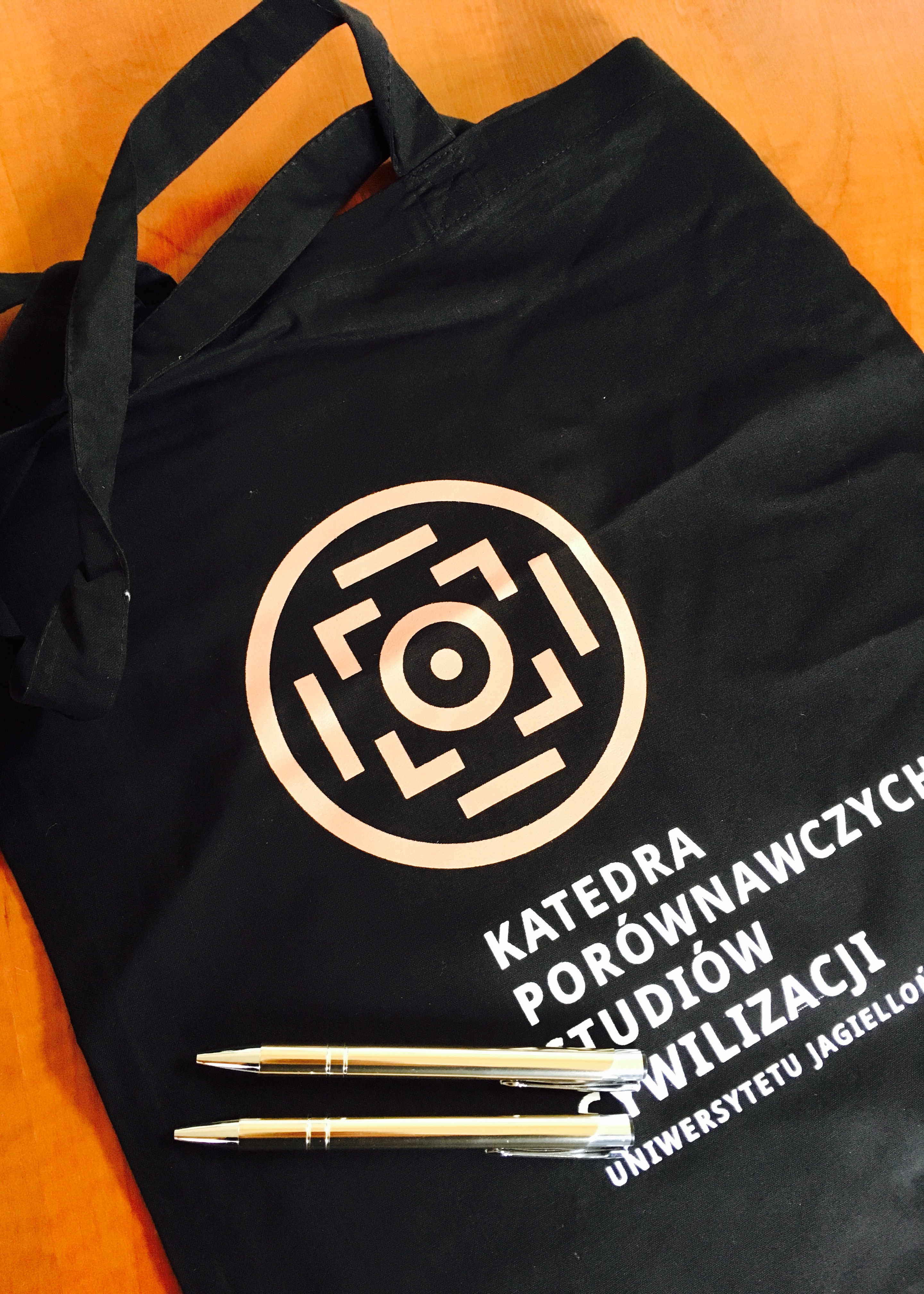 Zdjęcie przedstawia czarną torbę z logo KPSC i dwa złote długopisy z logo KPSC leżące na niej.