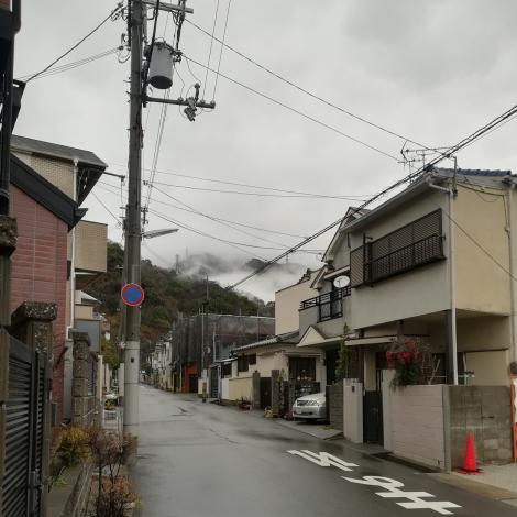 Zdjęcie nr 9 (13)
                                	                             Zdjęcie Tomasza Jakutowicza z pobytu w Japonii. Zdjęcie przedstawia ulicę, budynki mieszkalne, w tle wzgórza.
                            